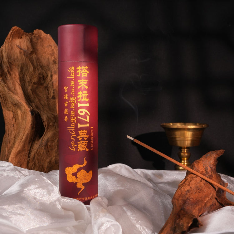 A cylindrical red incense stick container with gold Chinese text and a gold cloud graphic, standing on white fabric beside a burning incense stick, a brass bowl, and rustic wood pieces against a dark background.