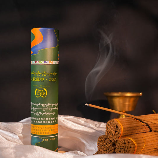 A cylindrical incense stick container labeled in Tibetan script stands upright on draped white fabric, with a single incense stick burning and several sticks beside it against a dark smoky background.