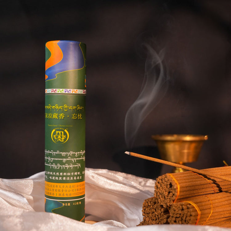 A cylindrical incense stick container labeled in Tibetan script stands upright on draped white fabric, with a single incense stick burning and several sticks beside it against a dark smoky background.