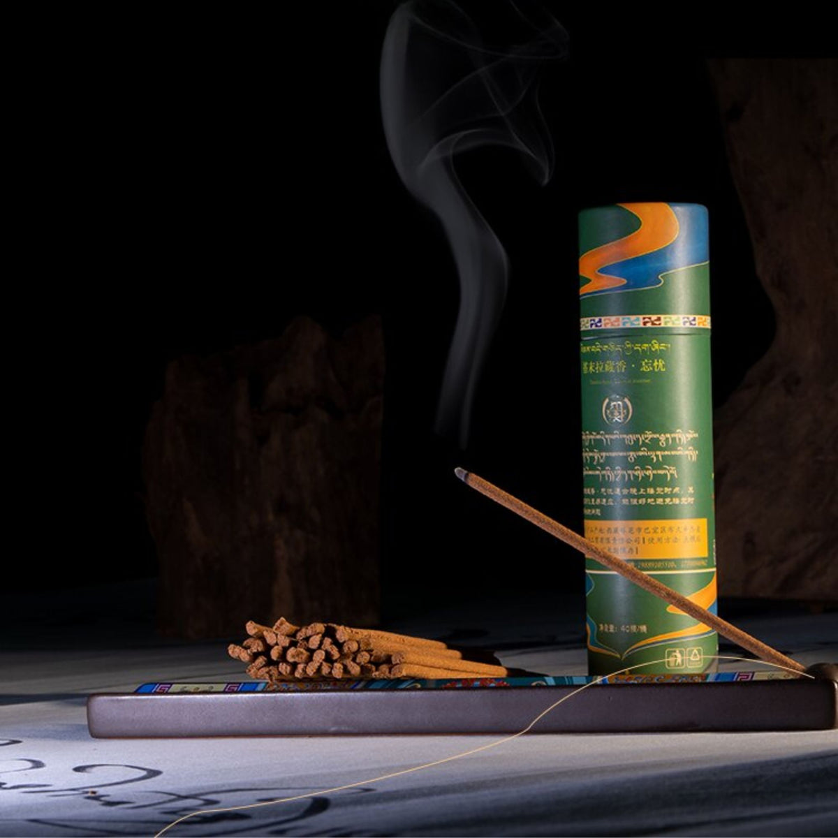 A cylindrical incense holder package in green, blue, and orange stands on a tray beside several incense sticks and a lit incense stick with visible smoke against a dark background.