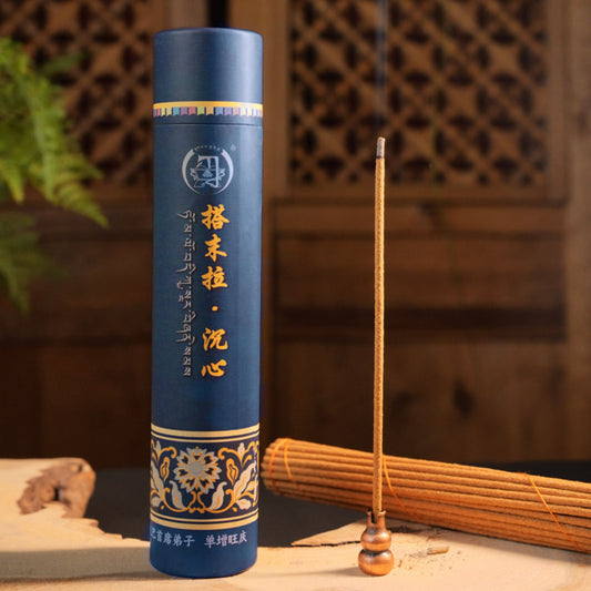A tall cylindrical blue incense holder or incense stick package stands on a tabletop beside a single lit incense stick and a rolled bundle of incense sticks. The background is a warm wooden interior with a lattice screen and soft blur.