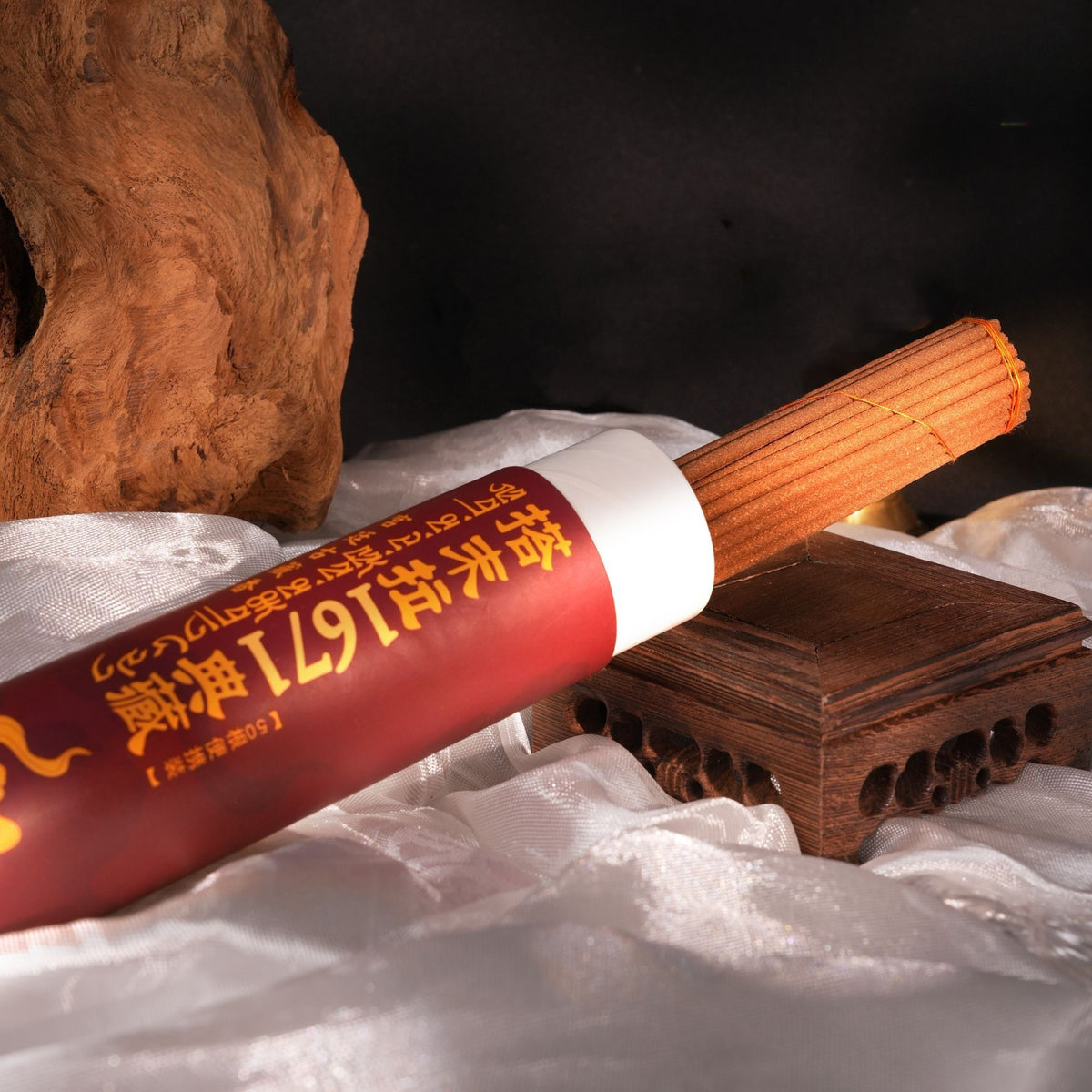 A red cylindrical incense stick holder with gold Chinese characters and a visible incense stick, styled on white fabric with a wooden prop and dark background.
