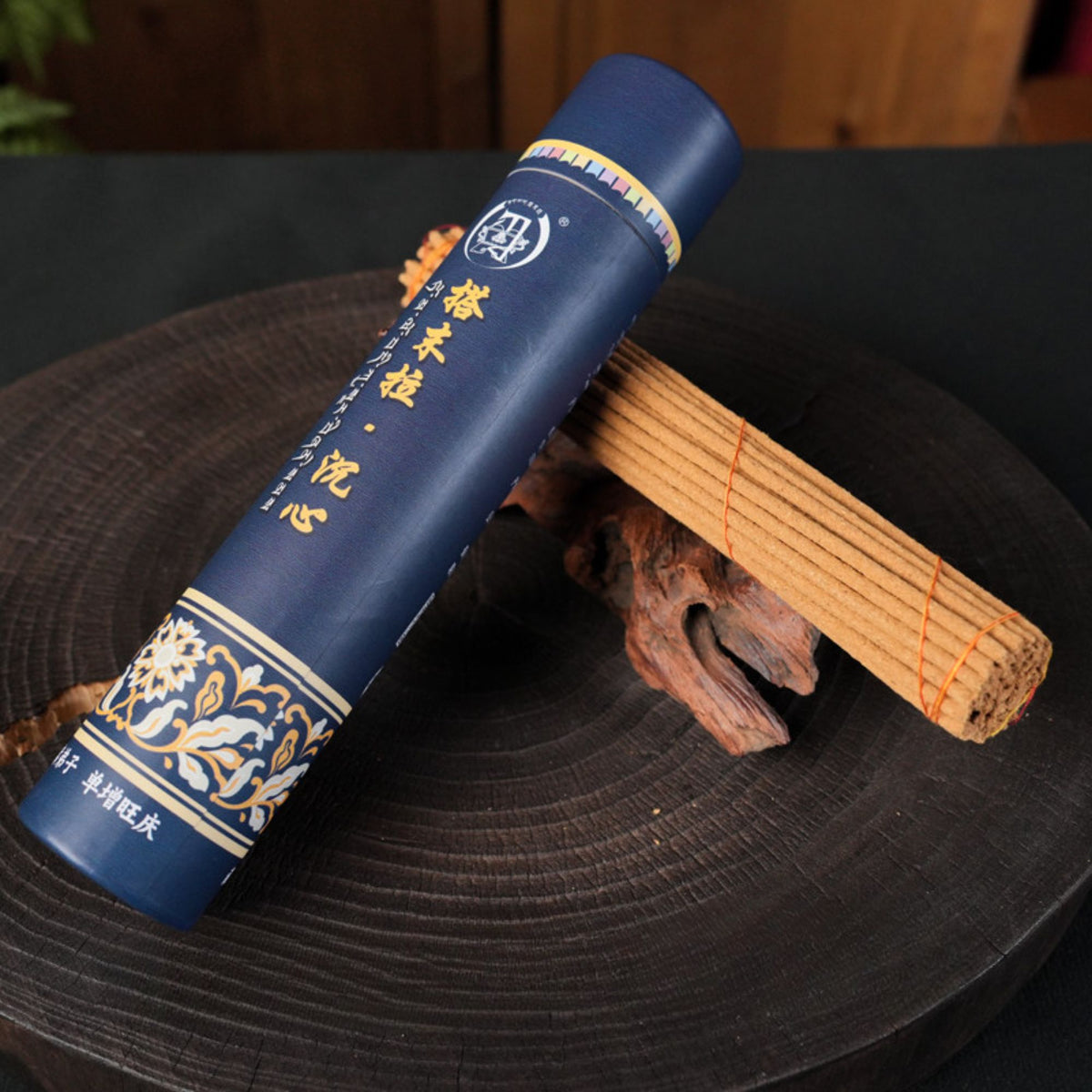 A dark blue cylindrical product package with gold Chinese characters and decorative floral patterns, placed diagonally on a round dark wooden tray with a bundle of incense sticks behind it.